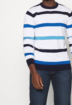 Person wearing white sweater with horizontal navy and blue stripes and dark blue jeans, standing against a plain light background.