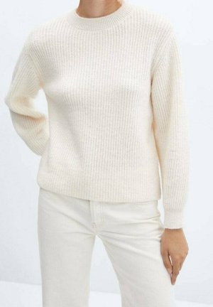 Femme portant un pull côtelé crème et un pantalon assorti de couleur claire, debout avec une main sur la hanche.