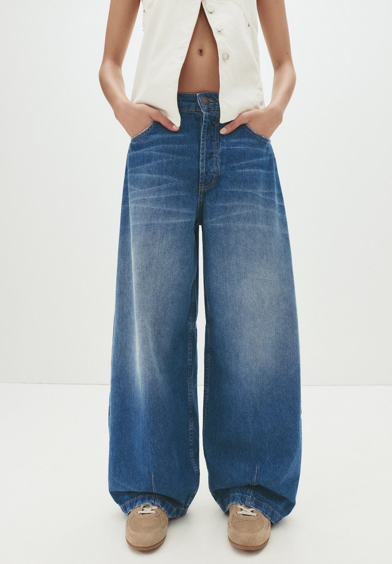 Personne portant un jean bleu à large jambe, des baskets beiges et un top sans manches blanc ouvert montrant un ventre nu, les mains dans les poches, sur fond blanc.