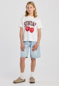 Camiseta gráfica blanca con el texto "SUNDAY" y estampados de cereza rojos, combinada con pantalones cortos de mezclilla azul claro y zuecos beige con una correa.