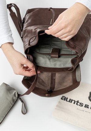 Manos abriendo una mochila de cuero marrón que muestra un forro interior a rayas, junto a un pequeño estuche a rayas y una bolsa de tela beige con texto.