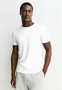 Balta īsroku T-krekls ar apaļu kakla izgriezumu, izgatavots no gludas auduma. Modelis valkā pelēkas šortus, kas atklāj tetovējumu uz rokas.