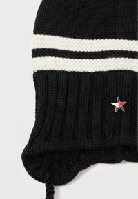 Bonnet en tricot noir avec des rayures horizontales blanches et une texture côtelée, arborant un petit accent étoilé rouge, blanc et bleu sur le côté.