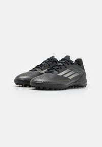 Botas de fútbol negras con parte superior sintética texturizada, que presentan tres franjas blancas y acentos dorados. Diseñadas para tracción con tacos de goma.