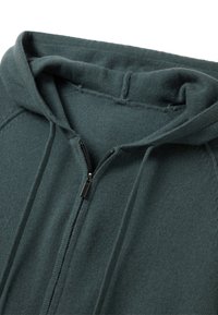 Felpa con zip di colore verde scuro, realizzata in un tessuto testurizzato, con cappuccio attaccato e zip nera. Cuciture visibili e vestibilità casual e rilassata.