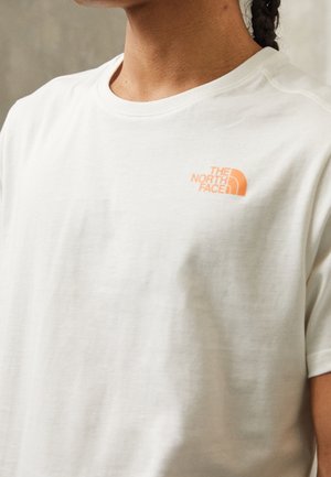 Osoba ubrana w biały T-shirt z okrągłym dekoltem z małym pomarańczowym logiem The North Face na górnej lewej stronie klatki piersiowej, włosy splecione z tyłu.