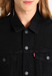 Osoba v černé džínové bundě Levi's s kovovými knoflíky a červenou visačkou Levi's, záběr z blízka od hrudníku k rtům.