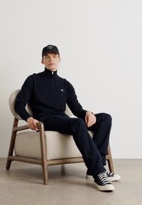 Black zip-up sweater and dark pants worn by a person seated in a wooden chair, paired with black and white sneakers and a black cap.