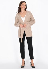 Beżowy kardigan z wiązanym w pasie, z perłowymi akcentami na ramionach, zestawiony z czarnymi, dopasowanymi spodniami i czarnymi szpilkami; luźny krój, miękka faktura.