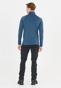 Pullover blu aderente a maniche lunghe con colletto alto, maniche raglan e tessuto elasticizzato. Indossato con pantaloni neri e stivali.