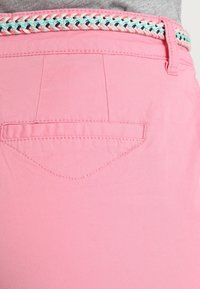 Rosa byxor har en slät tygtextur, med sömnadsdetaljer och en liten bakficka. Ett flätat bälte med turkosa och korallfärgade accenter fulländar looken.