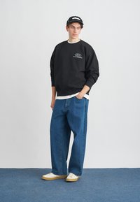 Črna jopa s kapuco z belim logotipom, nošena čez belo srajco. Modre široke kavbojke in bele superge z bež podplati. Črna kapa.