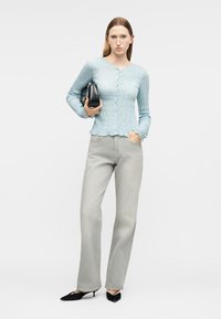 Γυναίκα που φοράει ένα ανοιχτό μπλε, υφασμάτινο τοπ με κουμπιά και ανοιχτόγρια ευθύγραμμα τζιν παντελόνια, κρατώντας μια μαύρη τσάντα, στέκεται μπροστά από ένα απλό φόντο.