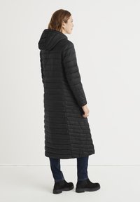 Polstret sort lang frakke med hætte, udstyret med et quiltet design og ribbet tekstur. Båret med mørke jeans og sorte ankelstøvler.