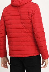 Röd vadderad jacka med horisontella quiltade sektioner, en huva och långa ärmar, tillverkad av lättviktigt tyg. Svarta byxor bärs.