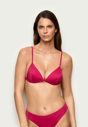 Mujer con cabello largo castaño, que lleva un bikini rojo sencillo con tirantes finos, de pie frente a un fondo claro y liso.