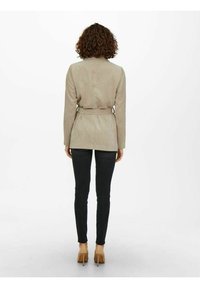 Blazer in corda beige con cintura a cravatta, maniche lunghe e vita aderente, abbinato a jeans skinny neri e tacchi alti nudi.