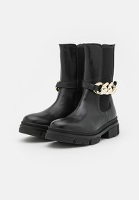 Botas de tobillo de cuero negro con suela gruesa, que cuentan con paneles laterales elásticos y un acento de cadena dorada en el lado exterior.