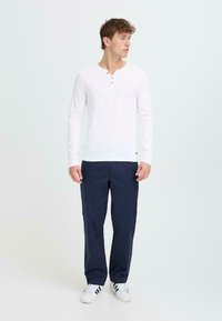 Bijela dugih rukava henley majica s tri gumba, uparena s hlačama tamnoplave boje. Model nosi bijele sportske tenisice.