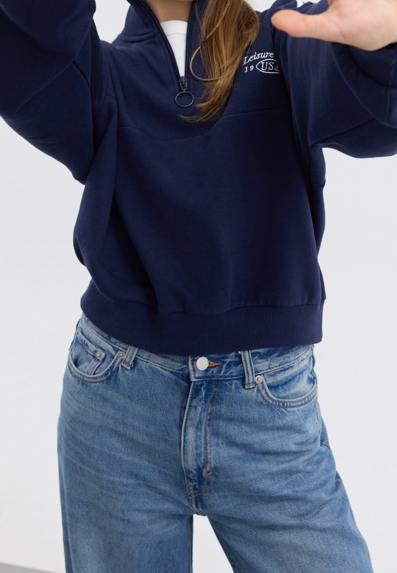 Personne portant un sweat-shirt à col montant bleu marine avec le logo "Leisure USA" et un jean taille haute bleu clair, tendant les bras en avant avec les mains levées.