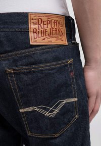 Jeans de mezclilla índigo con una etiqueta de cuero que presenta texto en rojo. El bolsillo trasero tiene un hilo beige en un patrón de ola. Diseño clásico de cinco bolsillos.