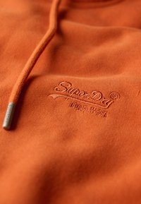 Sudadera con capucha de forro polar naranja que presenta un logo bordado de "SuperDry" y un detalle de metal en el cordón. Textura suave, diseño casual.