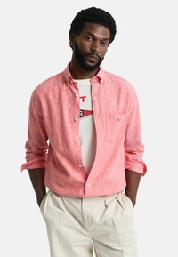Lichtroze overhemd met knopen van een gestructureerde stof, met een enkele borstzak, gedragen open over een wit T-shirt. Gepaard met beige geplisseerde broek.