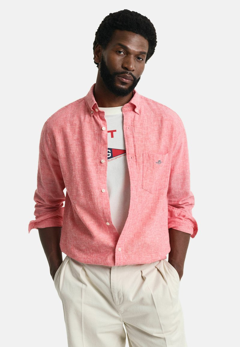 Lichtroze overhemd met knopen van een gestructureerde stof, met een enkele borstzak, gedragen open over een wit T-shirt. Gepaard met beige geplisseerde broek.