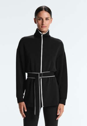 Žena u crnoj jakni na patentni zatvarač s bijelim obrubom i odgovarajućim remenom u struku, stoji ispred jednostavne svijetle pozadine.