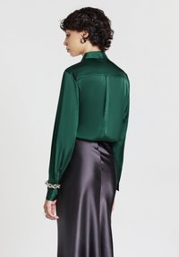 Blusa verde in satin con colletto strutturato, maniche lunghe e polsini con bottone, abbinata a una gonna nera in satin con cucitura laterale.