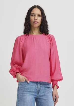 Blusa rosa con maniche a sbuffo, polsini arricciati e vestibilità rilassata. Realizzata in tessuto leggero con una texture liscia. Indossata con jeans blu.