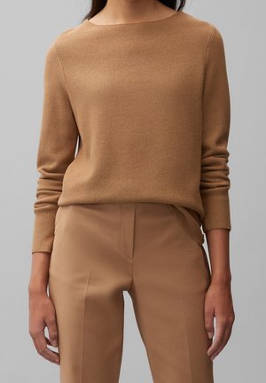 Vrouw draagt een getextureerde gebreide trui in camel en op maat gemaakte camelbroek terwijl ze staat tegen een effen grijze achtergrond.