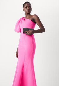 Fuchsiové, těsně přiléhající šaty s jedním ramínkem a velkým draped detailem. Modelka drží černou, zdobenou kabelku.