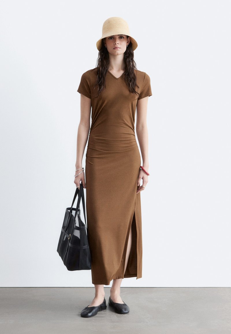 Femme portant une robe maxi marron à manches courtes avec une fente latérale, un chapeau beige, des ballerines noires et un grand sac cabas noir.