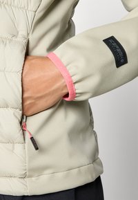 Beige takki, jossa on vaaleanpunaiset hihansuut ja musta logopatch. Varusteena näkyvä vetoketju ja joustavat hihansuut turvallista istuvuutta varten.
