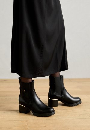 Des bottines noires en cuir présentent un talon bloc avec un accent doré et une semelle en caoutchouc texturée, complétées par des panneaux élastiques sur les côtés et une languette à l'arrière.