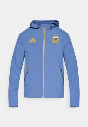 Hellblaue Jacke mit Kapuze, goldfarbenem Reißverschluss und Akzenten. Zeigt deutlich das Adidas-Logo und das Emblem des argentinischen Fußballverbands.