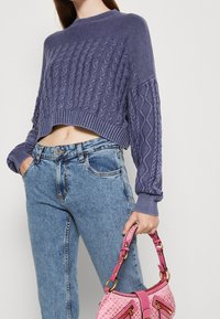 Fioletowy krótki sweter w warkocze z długimi rękawami, zestawiony z jasnoniebieskimi dżinsami z wysokim stanem. W ręku trzymana jest różowa torebka w wzory.