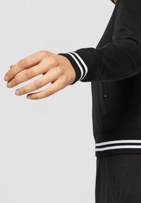 Bras droit tendu portant une veste noire avec des poignets et une ceinture à rayures blanches, sur un fond uni gris clair.