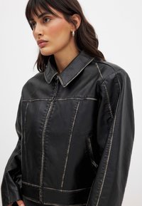 Moteris su tamsiais plaukais dėvi juodą odinį švarką su matomomis siūlėmis ir užtrauktuko kišene, šiek tiek žiūrinti į šoną prieš paprastą foną.