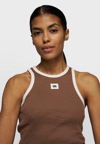 Braun gerippter Tanktop mit einem weißen Halsausschnitt und einem kleinen weißen Logo-Patch mit schwarzen Linien in der Mitte. Glatte Textur.