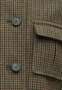 Brązowo-zielona tkanina w wzór houndstooth, z ciemnymi guzikami, klapką kieszeni oraz teksturalnymi detalami powierzchni.