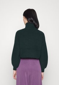 Maglione a collo alto a coste verde scuro con maniche a campana, di lunghezza cropped. Indossato con pantaloni viola a vita alta. Design semplice e testurizzato.
