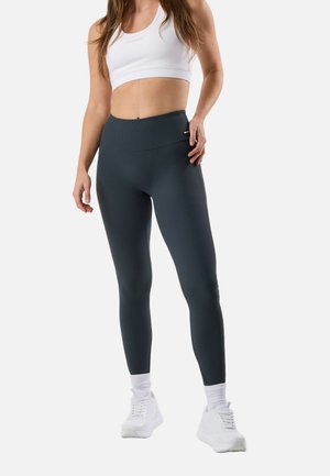 Frau trägt schwarze, voll lange Leggings, weißen Sport-BH, weiße Socken und weiße Turnschuhe, steht vor weißem Hintergrund.