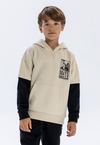 Beige hoodtröja med ett svart långärmat lager under, med en framficka och ett grafiskt tryck märkt "SKATE SKILLZ." Mjuk textur, avslappnad passform.