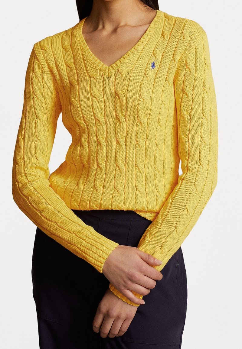 Femme portant un pull jaune en maille torsadée à col en V avec manches longues, associé à un pantalon sombre, les mains doucement croisées à la taille.