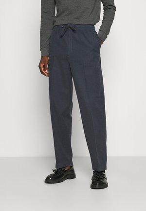 Mężczyzna ubrany w granatowe spodnie ze sznurkiem, ciemnoszary długi rękaw oraz czarne mokasyny z frędzlami stojący na jednolitym tle.