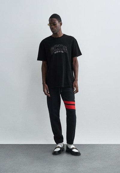 Juodas GCDS marškinėlis su siuvinėtų logotipu, derinamas su tamsiomis kelnėmis, turinčiomis raudonas juosteles. Avalynė - baltos loafer'iai su juodais akcentais.