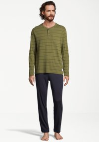 Mies seisoo paljain jaloin, päällä oliivinvihreä pitkähihainen henley-paita, jossa on ohuita raitoja, sekä mustat rentoa istuvuutta omaavat housut valkoista taustaa vasten.