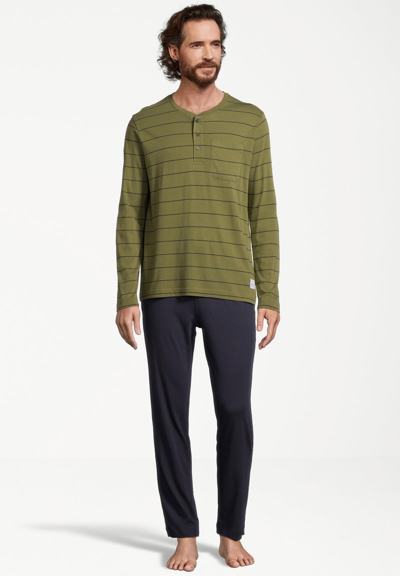 Mies seisoo paljain jaloin, päällä oliivinvihreä pitkähihainen henley-paita, jossa on ohuita raitoja, sekä mustat rentoa istuvuutta omaavat housut valkoista taustaa vasten.